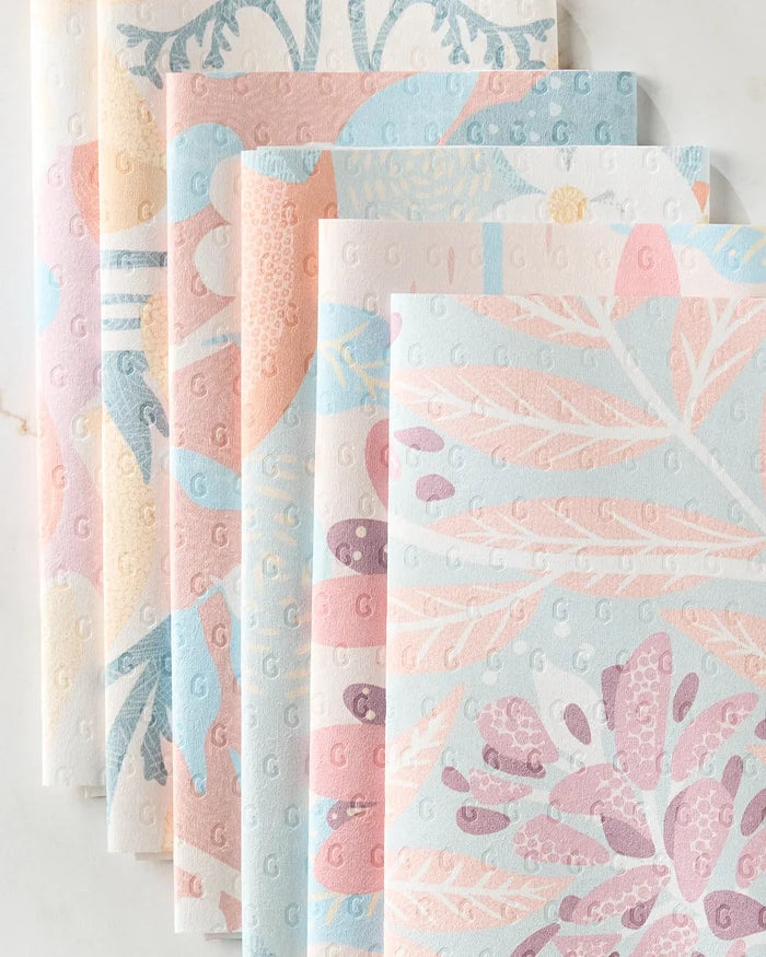 Six folded State Flowers paper napkins by Julianne Haness are displayed on a white surface, featuring pastel floral and botanical patterns in pink, blue, purple, and cream with embossed dots and subtle decorative designs.