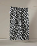 The Geometry Static kitchen towel, featuring a black and white pixelated checkered pattern, hangs from a string against a plain light background.