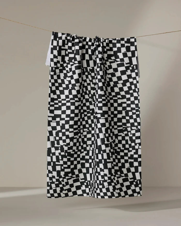 The Geometry Static kitchen towel, featuring a black and white pixelated checkered pattern, hangs from a string against a plain light background.