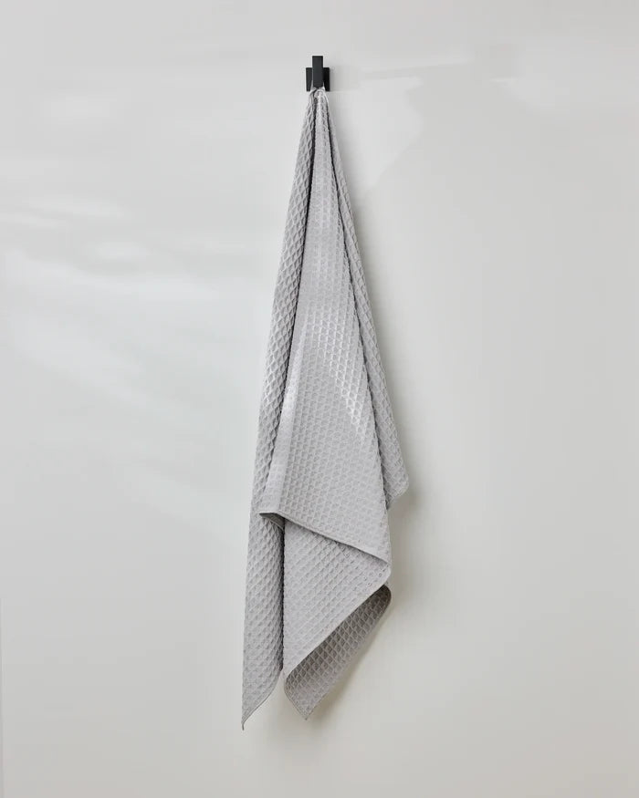 The GEOMETRY Stone waffle bath towel in light gray hangs neatly on a black hook against a plain white wall, adding style and comfort.