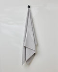 The GEOMETRY Stone waffle bath towel in light gray hangs neatly on a black hook against a plain white wall, adding style and comfort.