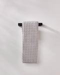 A light gray, waffle-textured GEOMETRY "Stone" luxury bath towel is neatly draped over a sleek black towel rack mounted on a plain, light-colored wall.