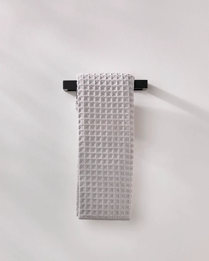 A light gray, waffle-textured GEOMETRY "Stone" luxury bath towel is neatly draped over a sleek black towel rack mounted on a plain, light-colored wall.