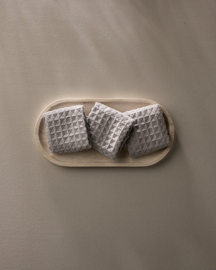 A wooden tray displays three neatly folded GEOMETRY Stone gray waffle washcloths, arranged in a row against a plain light beige background.