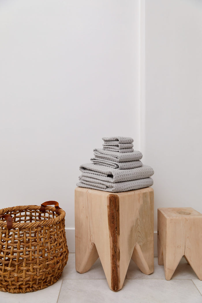 A stack of GEOMETRY Stone luxury towels sits on a light wooden stool beside a woven basket and a smaller wooden stool, set against a white wall and tiled floor.