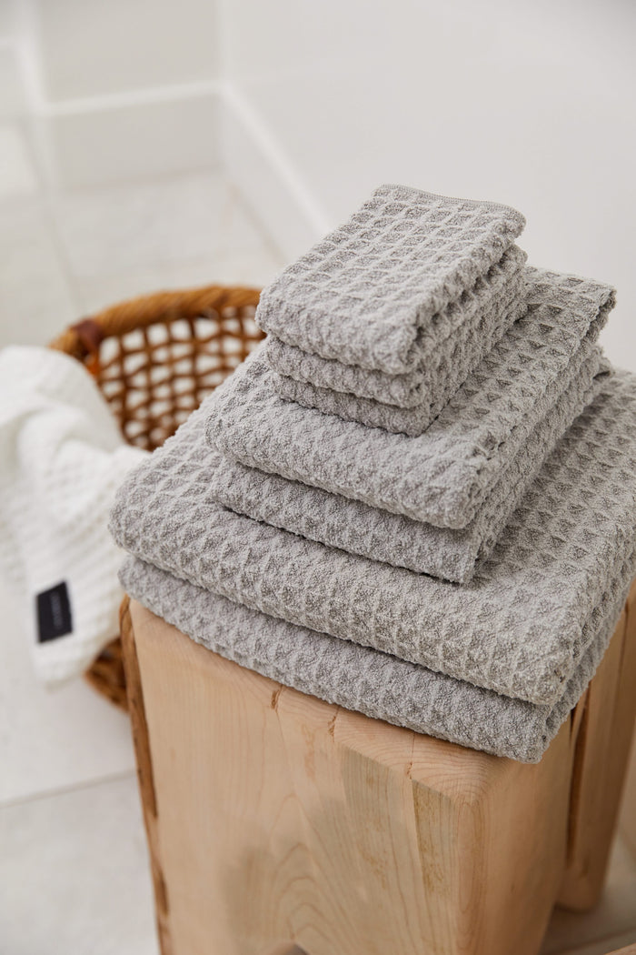 A neatly folded stack of GEOMETRY Stone ultra-absorbent waffle-textured towels sits on a wooden stool, with a wicker basket and a white Waffle Hand Towel in the background on a light tiled floor.