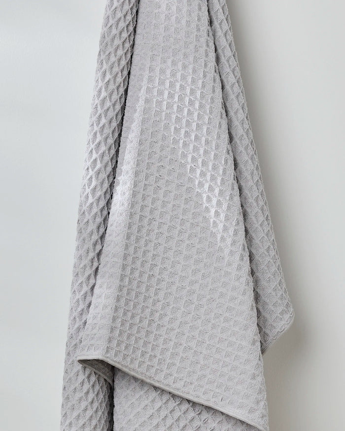 The Stone waffle bath towel by GEOMETRY, in gray with a grid-like texture, hangs in soft folds on a light wall, bringing style and comfort to your space.