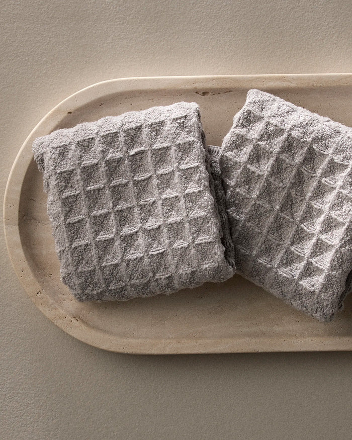 Two folded Stone gray waffle washcloths by GEOMETRY are placed side by side on an oval beige tray against a neutral background.