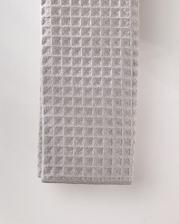 A GEOMETRY Stone hand towel in light gray, featuring a textured waffle pattern, hangs against a pale wall.