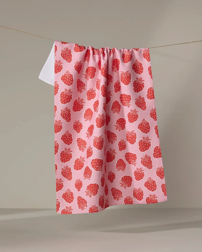 The Strawberry Fields tea towel by Charlie Rowan Designs, featuring a pink, ultra-absorbent fabric and red strawberry pattern, hangs on a clothesline against a plain background.