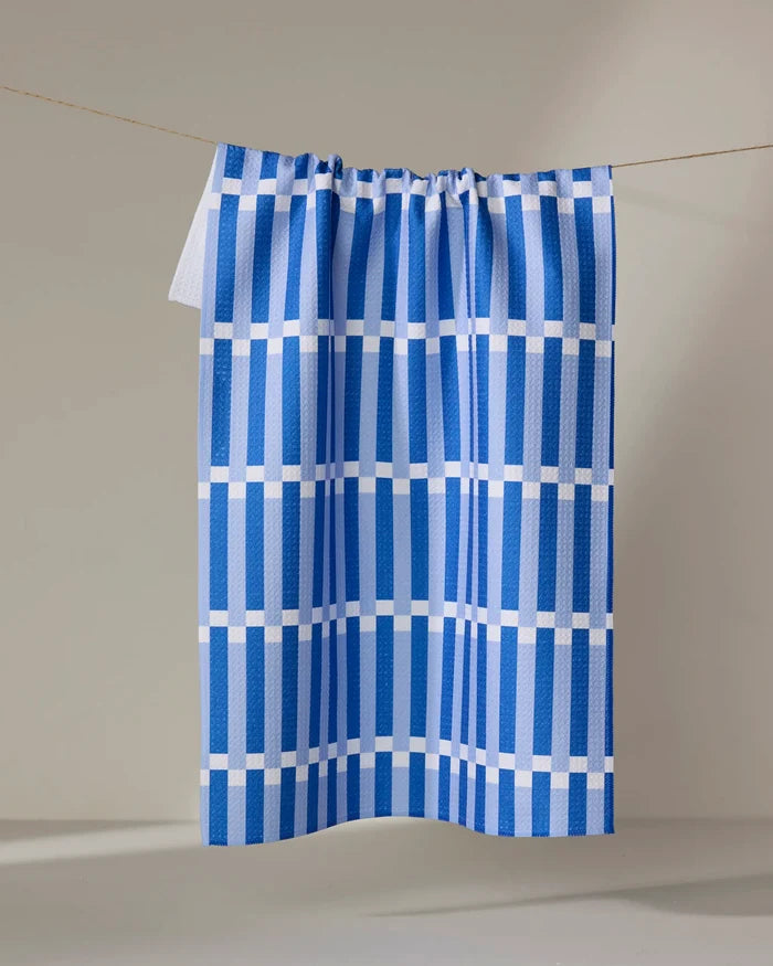 The Buy Guide’s Stripe Block - Blues is an absorbent kitchen towel made from recycled materials, featuring blue and white grid stripes. It hangs on a clothesline against a light background.