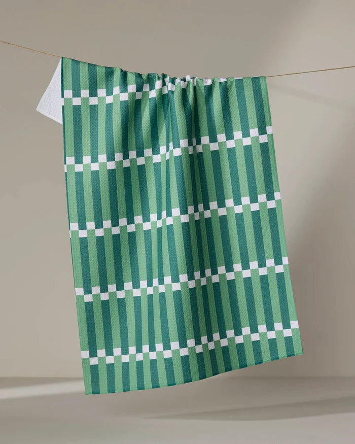 The Stripe Block - Greens tea towel by The Buy Guide hangs on a line against a neutral background, showcasing its green and white geometric pattern with vertical stripes and evenly spaced horizontal white lines.