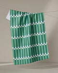 The Stripe Block - Greens tea towel by The Buy Guide hangs on a line against a neutral background, showcasing its green and white geometric pattern with vertical stripes and evenly spaced horizontal white lines.