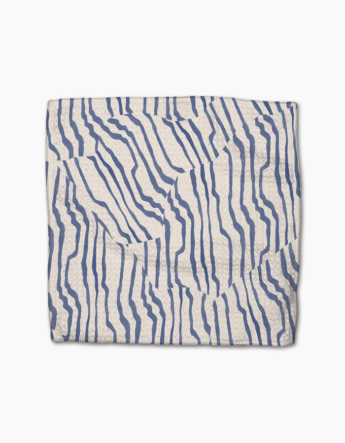 The Geometry Stripe Funk Grounds is a folded, cream-colored blanket made from recycled materials, featuring abstract wavy blue lines on a white background.