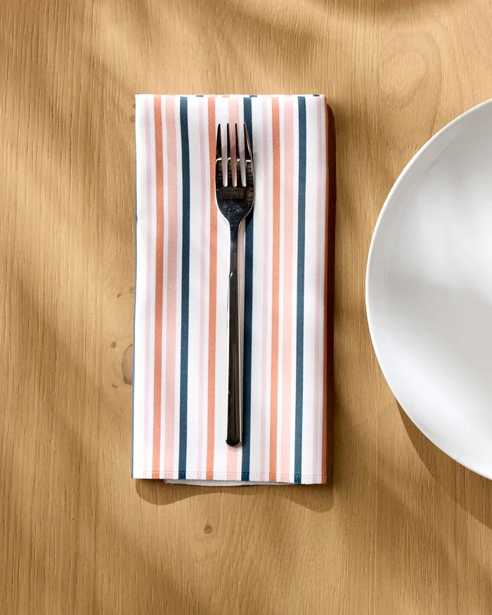 A metal fork rests on a neatly folded Striped Harvest napkin by Charlie Rowan Designs, featuring pink, white, and gray stripes, beside a white ceramic plate on a light wooden table.