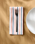 A metal fork rests on a neatly folded Striped Harvest napkin by Charlie Rowan Designs, featuring pink, white, and gray stripes, beside a white ceramic plate on a light wooden table.