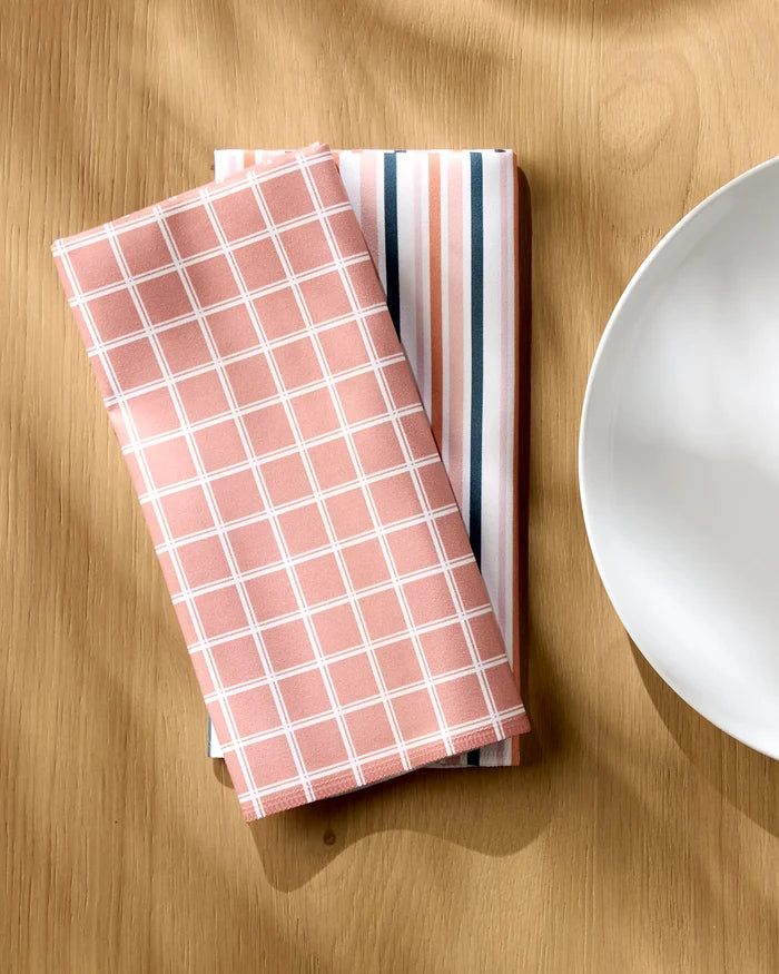 Two neatly folded Striped Harvest napkins by Charlie Rowan Designs are placed side by side on a wooden table next to a white ceramic plate.