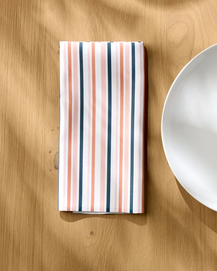 The Striped Harvest reusable dinner napkin by Charlie Rowan Designs, featuring pink, white, and dark gray stripes, is neatly folded on a light wooden table beside a white plate with sunlight casting gentle shadows.