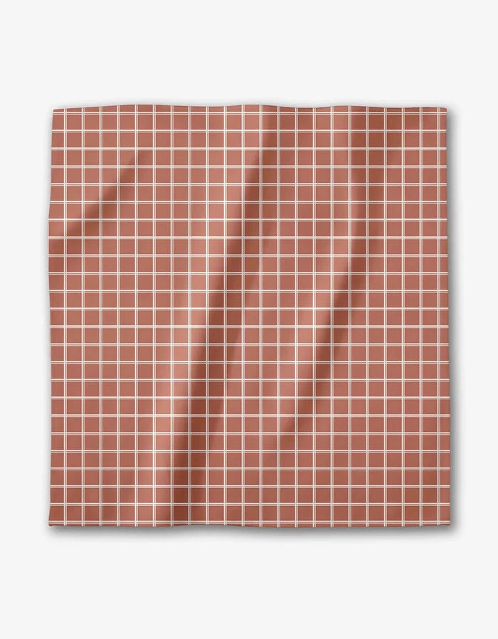 The Striped Harvest dinner napkin by Charlie Rowan Designs features a square Geosuede fabric with a terracotta background, white grid pattern, and gentle folds and creases.