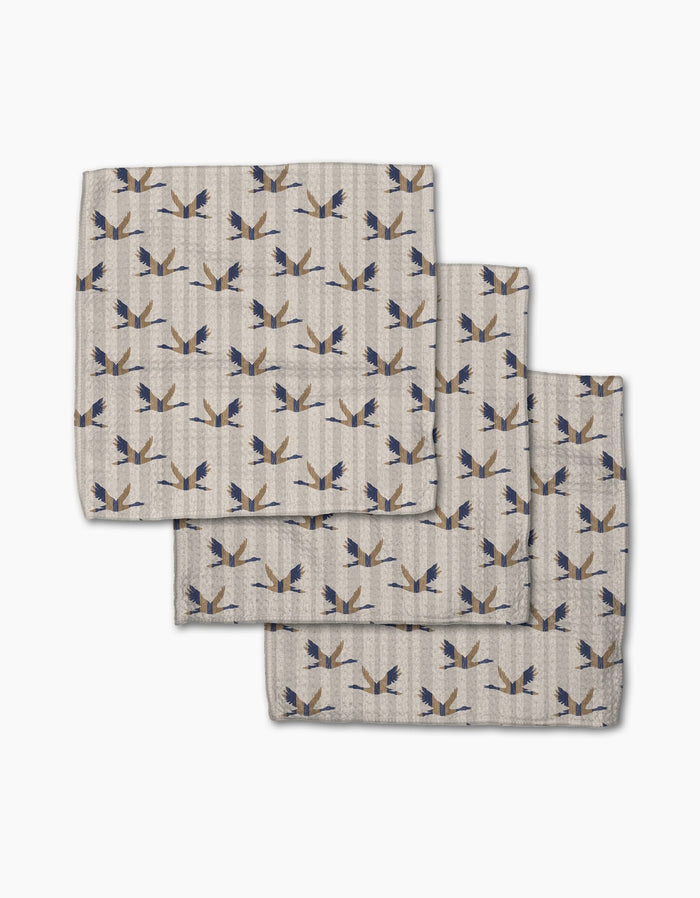 Three folded Striped Nautical Birds napkins by Geometry, featuring a blue and gold bird pattern on beige Geoweave fabric, are overlapped on a white background.