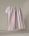 The Stripes For Days tea towel by Charlie Rowan Designs, featuring ultra-absorbent fabric and vertical pink, white, and dark green stripes, hangs on a clothesline against a plain beige background.