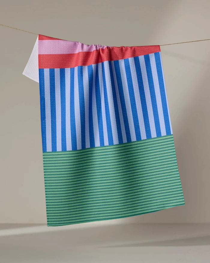 The Buy Guide’s Stripes on Stripes recycled kitchen towel, featuring a red and pink top border, blue and white vertical stripes, and green bottom stripes, hangs on a line against a neutral background.