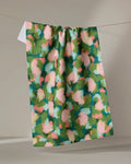 The "Stroke of Genius" tea towel by Shelley Smith, featuring an abstract floral design in green, pink, peach, and blue, hangs on a line against a neutral background.