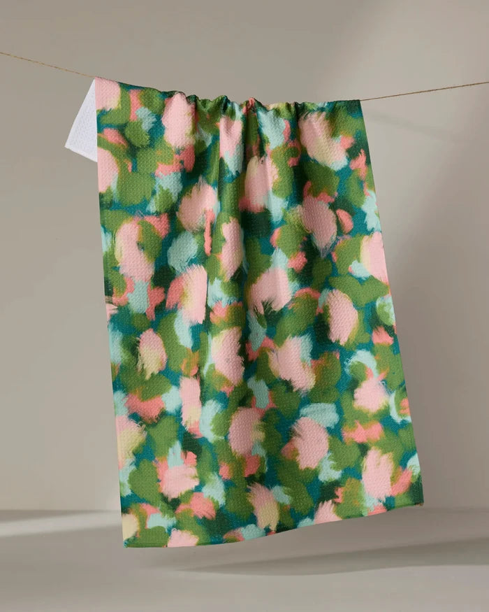 The "Stroke of Genius" tea towel by Shelley Smith, featuring an abstract floral design in green, pink, peach, and blue, hangs on a line against a neutral background.