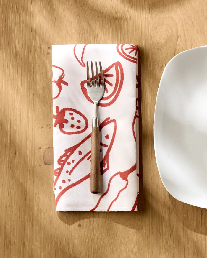 The Geometry "Summer Air" reusable cloth napkin with red fruit and vegetable designs is shown under a fork with a wooden handle, beside a white plate on a light wooden table.