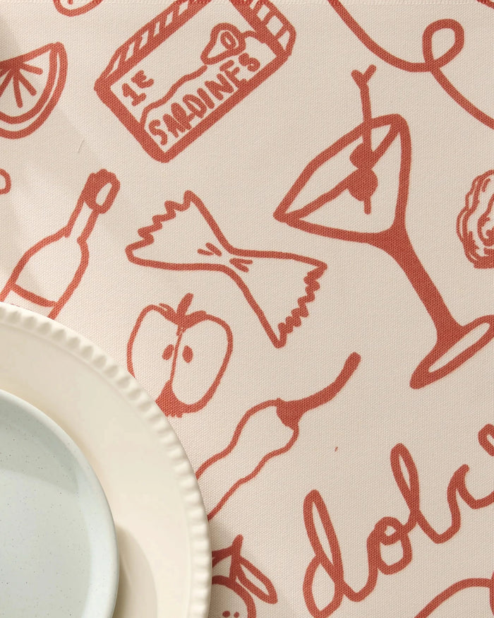 A close-up of Geometrys Summer Air tablecloth, featuring red food-themed illustrations. A white plate rests on recycled placemats in the bottom left corner.
