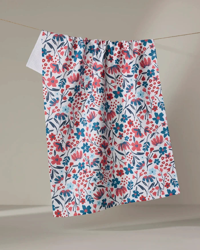 The Summer Bloom kitchen towel by Hufton Studio, crafted from recycled materials, features a colorful red, pink, and blue floral pattern on white and hangs on a string against a neutral background.