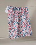 The Summer Bloom kitchen towel by Hufton Studio, crafted from recycled materials, features a colorful red, pink, and blue floral pattern on white and hangs on a string against a neutral background.