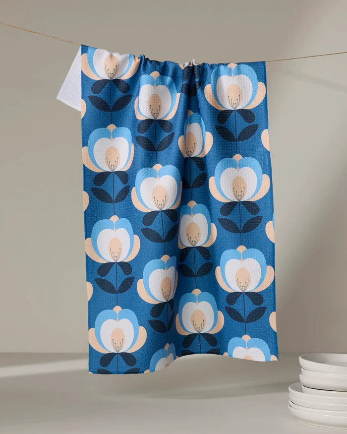 The Blissful Flamingo "Summer Florals" tea towel, featuring a blue and beige floral pattern, hangs on a line with white plates stacked nearby against a neutral backdrop.