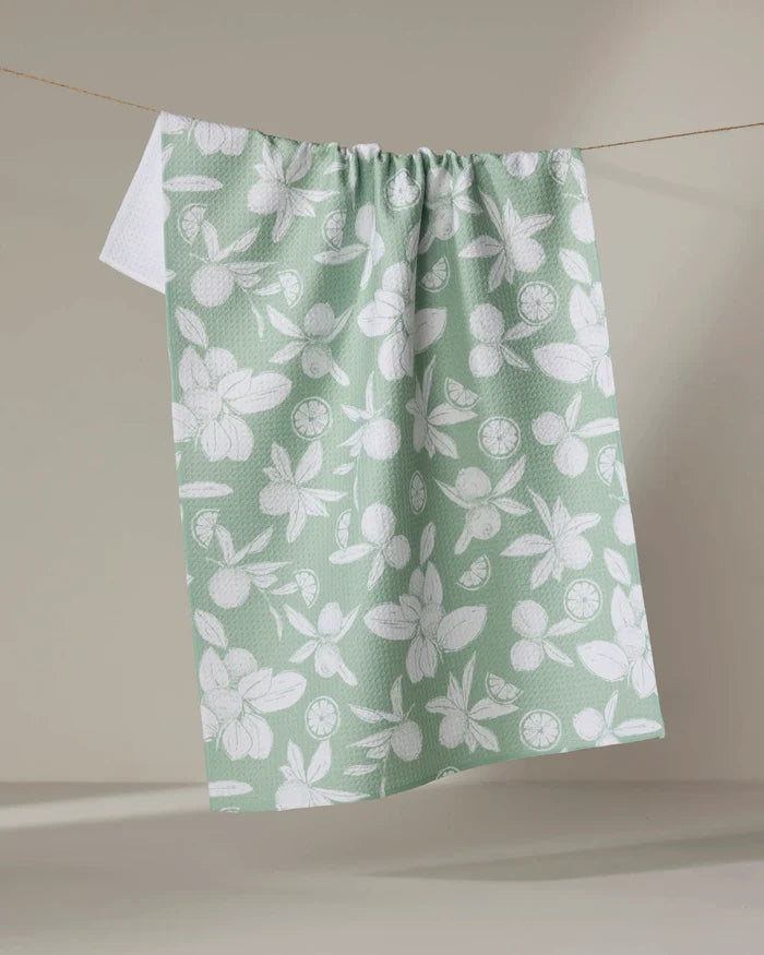 The Summer Limes towel by Hufton Studio is a quick-drying green tea towel made from recycled materials, featuring a white citrus fruit and leaf pattern, hanging on a line against a neutral background.