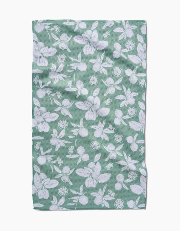 A green rectangular Summer Limes tea towel by Hufton Studio, made from recycled materials, features a white floral and citrus fruit pattern and is shown flat with slight wrinkles.
