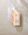 Seven folded Summer Picnic Dreams reusable towels by Letters By Shells, featuring pastel blue, purple, and yellow checkered patterns with fruit and animal designs, are neatly arranged as an eco-friendly kitchen option.