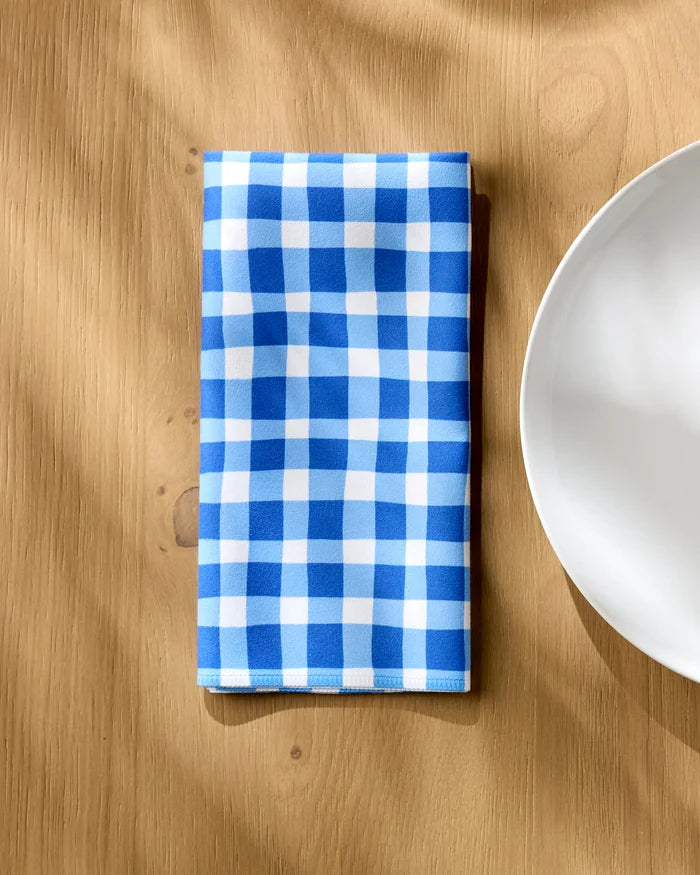 A Danelys Sidron Summer Plaid blue and white checkered eco-friendly napkin is neatly folded on a light wood table beside a white plate.
