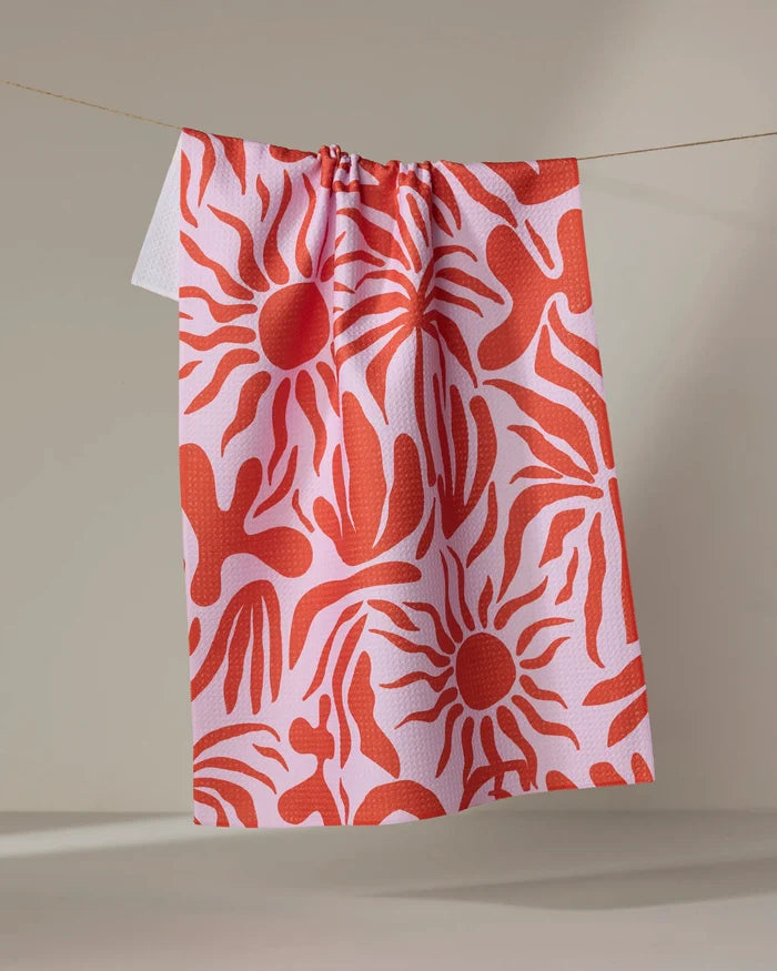 The "Summer Sunset" recycled kitchen towel by Marina Solodka, featuring bold red sunburst and hand-like patterns on pink, hangs on a string against a beige background.