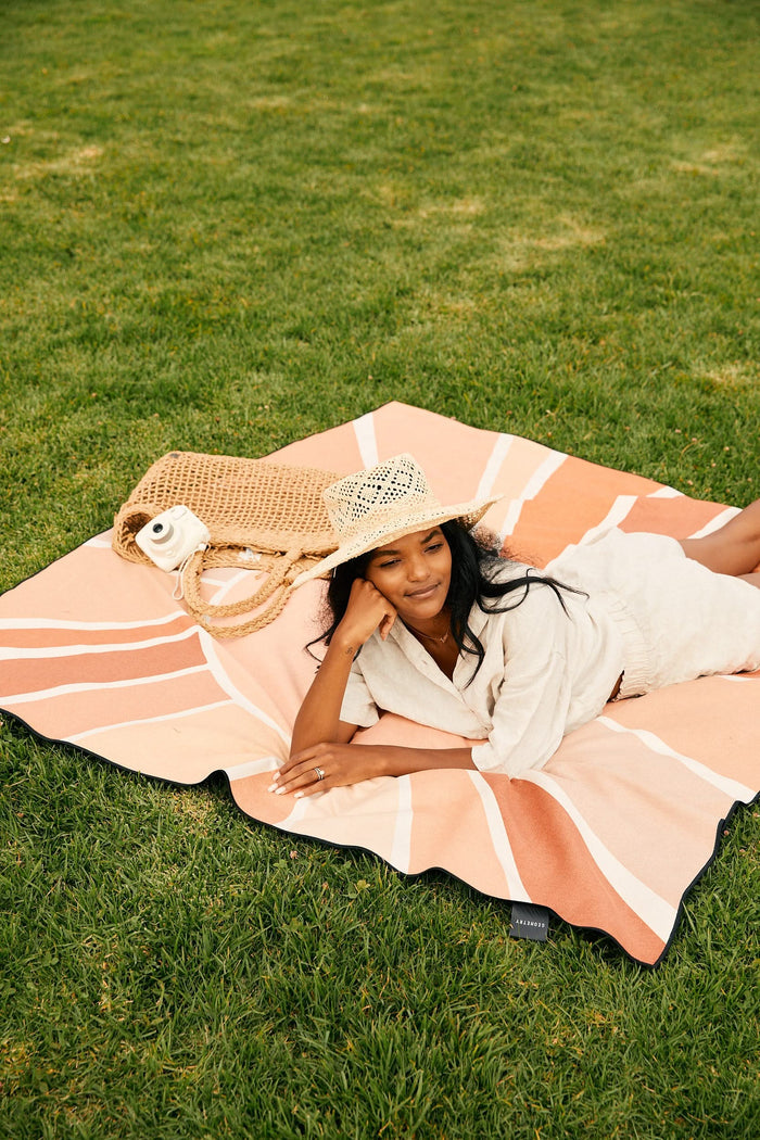 A woman in a light outfit and straw hat relaxes on the grass atop the GEOMETRY Sun And Stripes beach blanket, made from recycled materials, with a woven bag and water bottle beside her. She smiles slightly, enjoying her day outdoors.