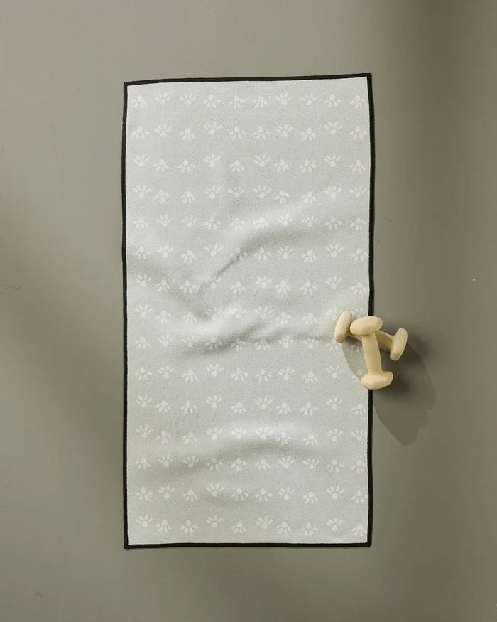 A single small dumbbell rests beside an ultra-absorbent towel on the "Sun Bursts" yoga mat by Anee Shah, featuring light gray with subtle white floral designs, placed on a smooth gray surface.