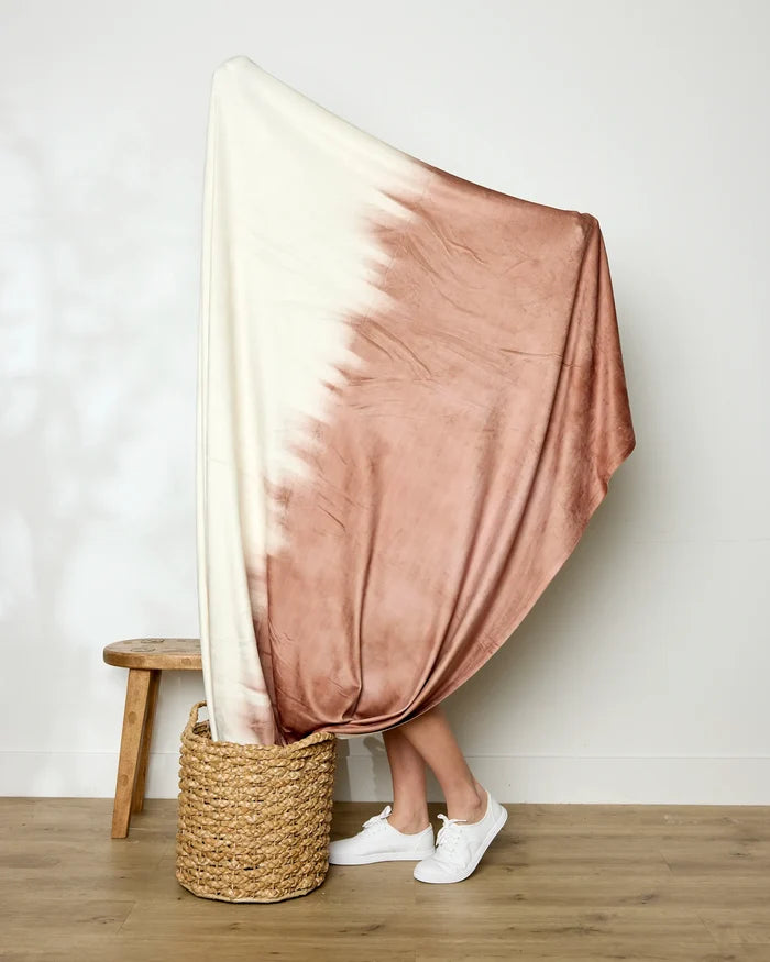 A person holds up the large Geometry Sundae Butter Blanket, featuring a white and blush pink tie-dye pattern. Only their lower legs and white sneakers are visible, with a woven basket and wooden stool nearby, showcasing its silky softness.