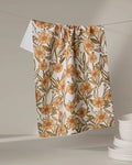 The Sunflower Fields tea towel by Janna Sue Design, featuring an ultra-absorbent orange and yellow floral print, hangs on a string near stacked white plates against a neutral background.