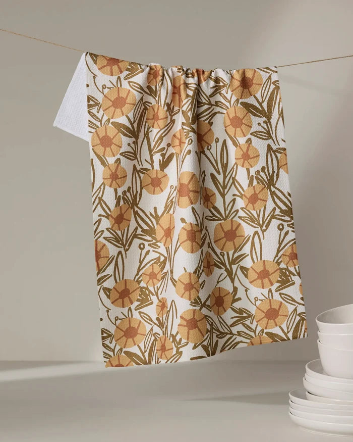 The Sunflower Fields tea towel by Janna Sue Design, featuring an ultra-absorbent orange and yellow floral print, hangs on a string near stacked white plates against a neutral background.