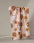 The Elisabeth Wing "Sunflowers" tea towel features pink fabric adorned with yellow and white sunflowers and daisies, hanging on a clothesline against a neutral backdrop to bring charm to your kitchen décor.