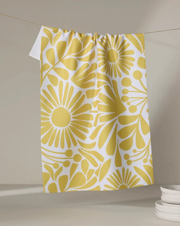 The Sunlit Blooms kitchen towel by Lisa Nelson, made from recycled materials, showcases a bold yellow floral design and hangs on a clothesline. White plates are stacked in the lower right against a neutral backdrop.