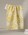 The Sunlit Blooms kitchen towel by Lisa Nelson, made from recycled materials, showcases a bold yellow floral design and hangs on a clothesline. White plates are stacked in the lower right against a neutral backdrop.