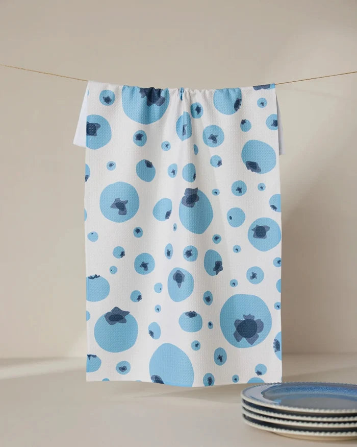 The GEOMETRY Sunny Blueberries white tea towel, crafted from absorbent Geoweave fabric and featuring a blue and dark blue blueberry pattern, hangs on a thin line. Blue-rimmed plates are partially seen in the lower right against a neutral background.