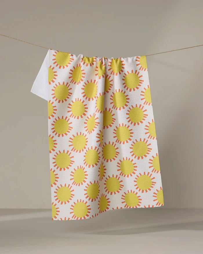 The Lizzie Clark "Sunny Day" tea towel, a quick-drying white cloth with yellow suns and orange rays, hangs on a clothesline against a neutral background.