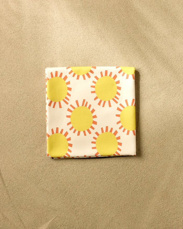 The Lizzie Clark Sunny Day Micro-loop Beach Blanket, quick-drying and comfortable, displays a bold yellow sun and orange ray pattern. Shown folded on a beige surface, it blends vibrant style with practical use.