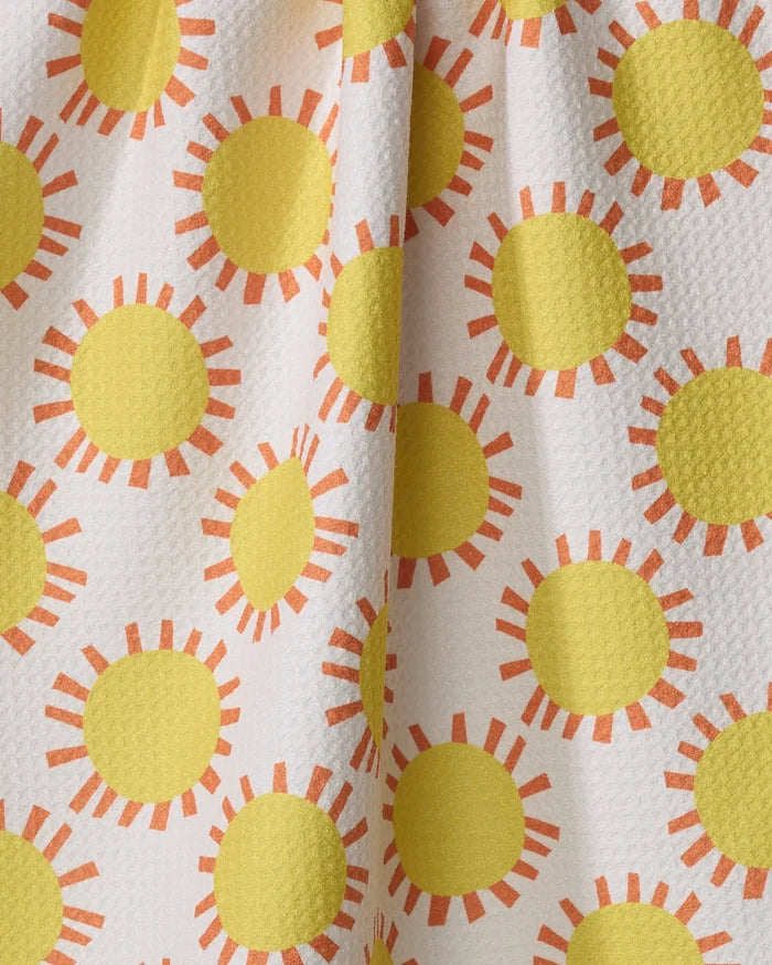 Lizzie Clark’s Sunny Day tea towel features a textured white fabric with yellow sun patterns and orange rays. Made from quick-drying recycled materials, its gathered construction creates soft folds and playful shadows.